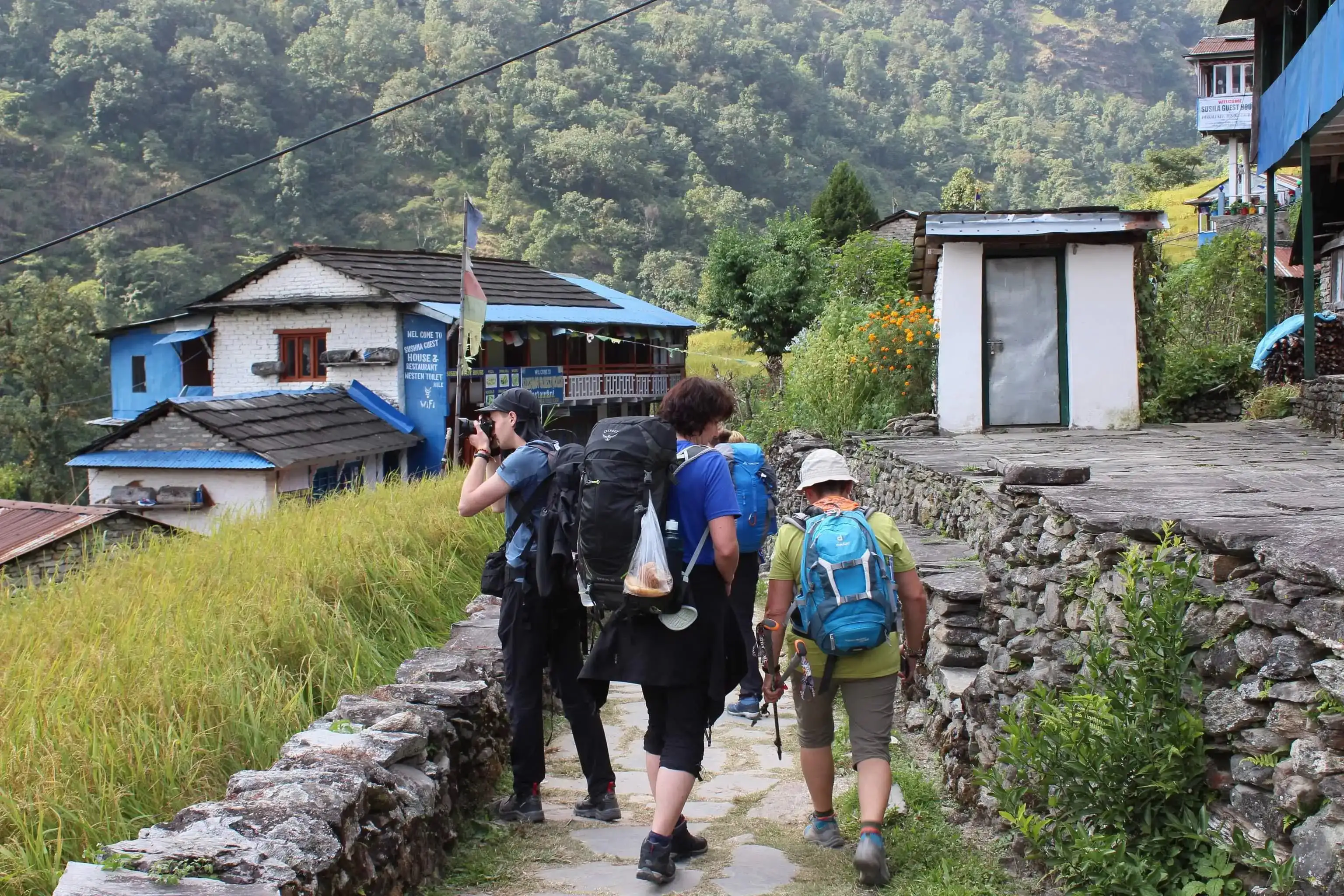 Hikers with backpacks walking through a stone path near village houses surrounded by green mountains.