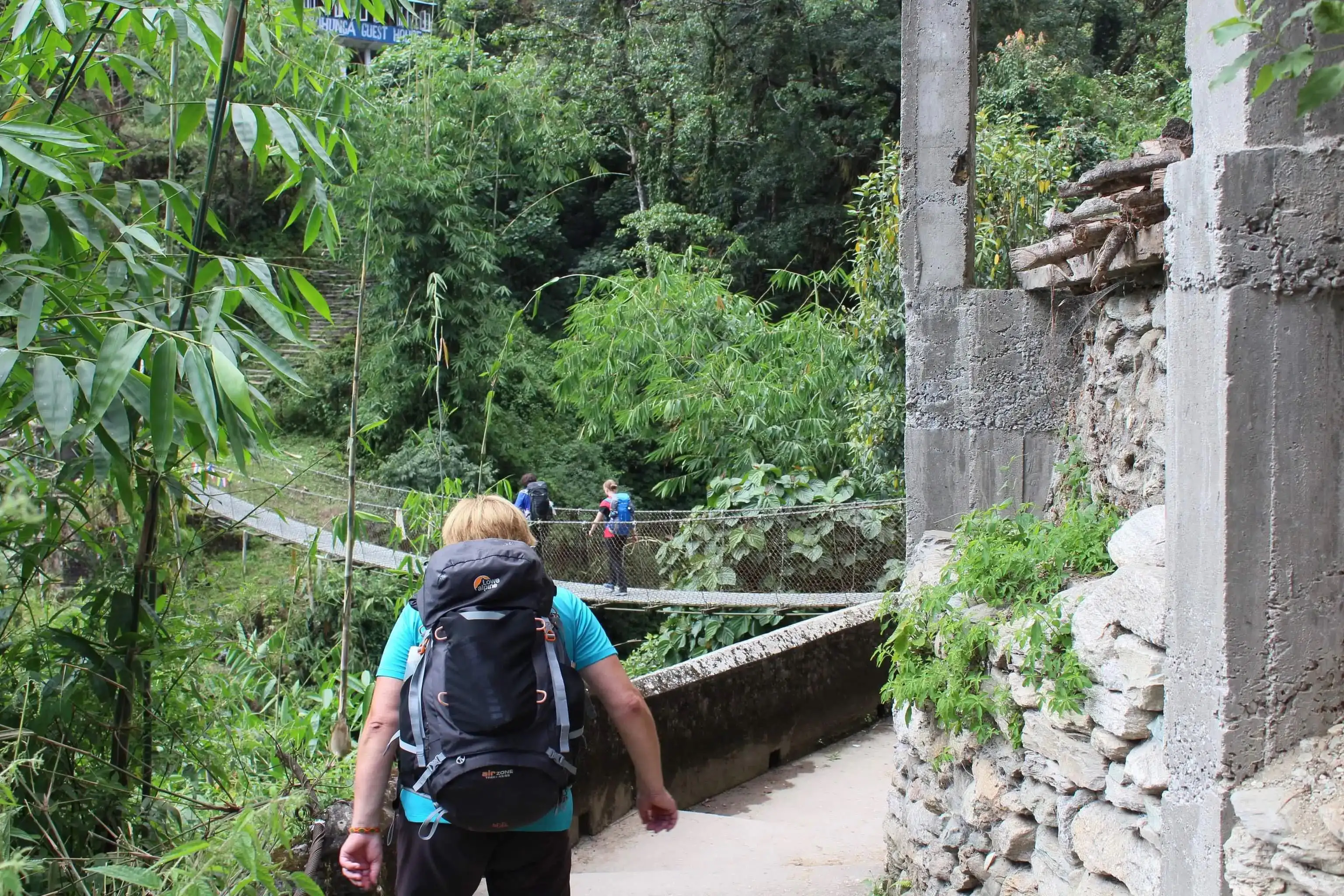Hiker with backpack walking towards a suspension bridge surrounded by dense green forest.